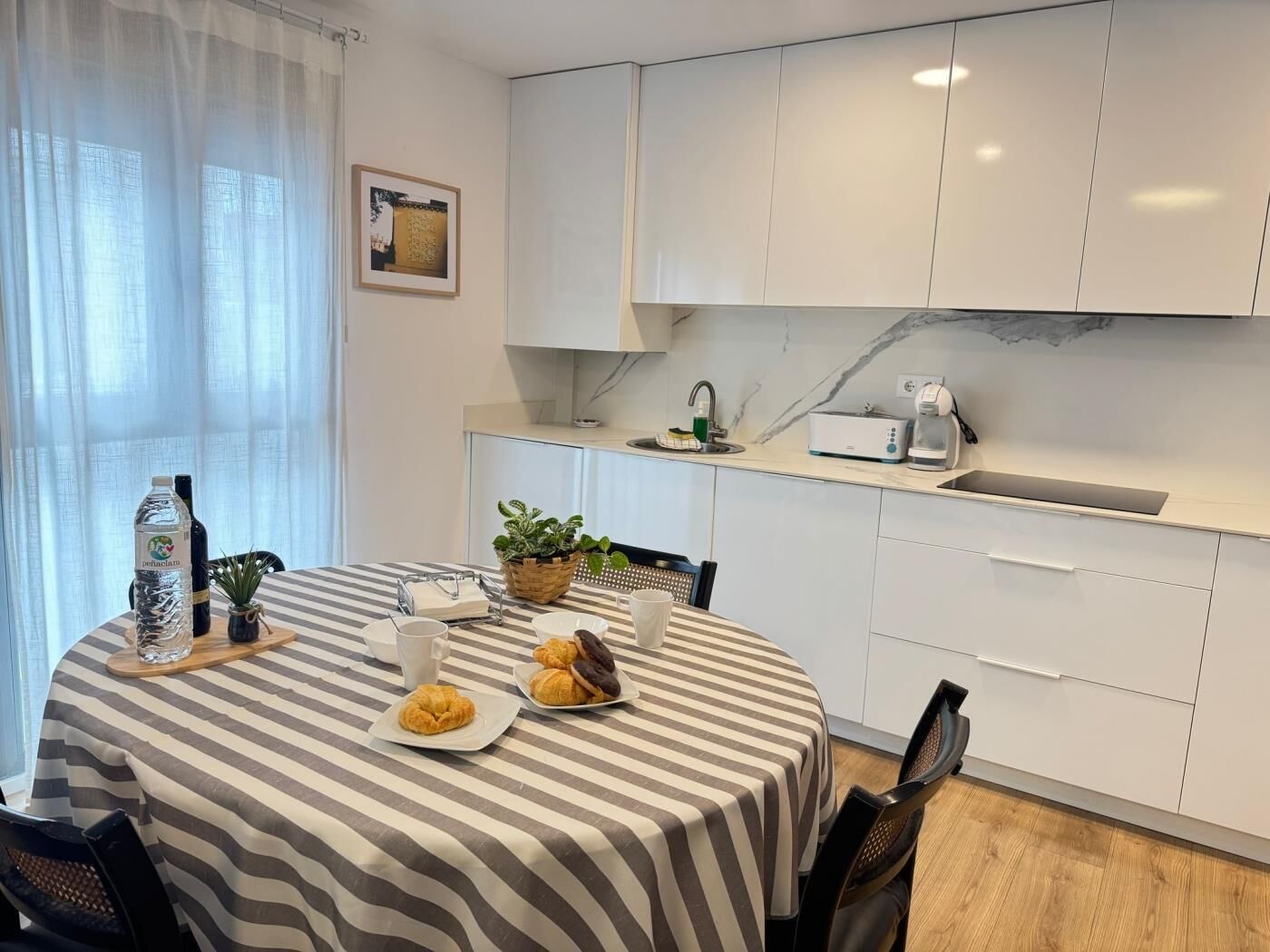 A modern kitchen with white cabinets, a striped tablecloth, a coffee maker, and a potted plant.