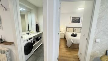 Habitación de hotel con cama, mesita de noche, espejo y baño con lavamanos y lavadora.