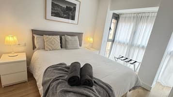 Una cama bien hecha con una toalla enrollada, dos lámparas de noche, un cuadro enmarcado encima de la cama y una ventana con cortinas.