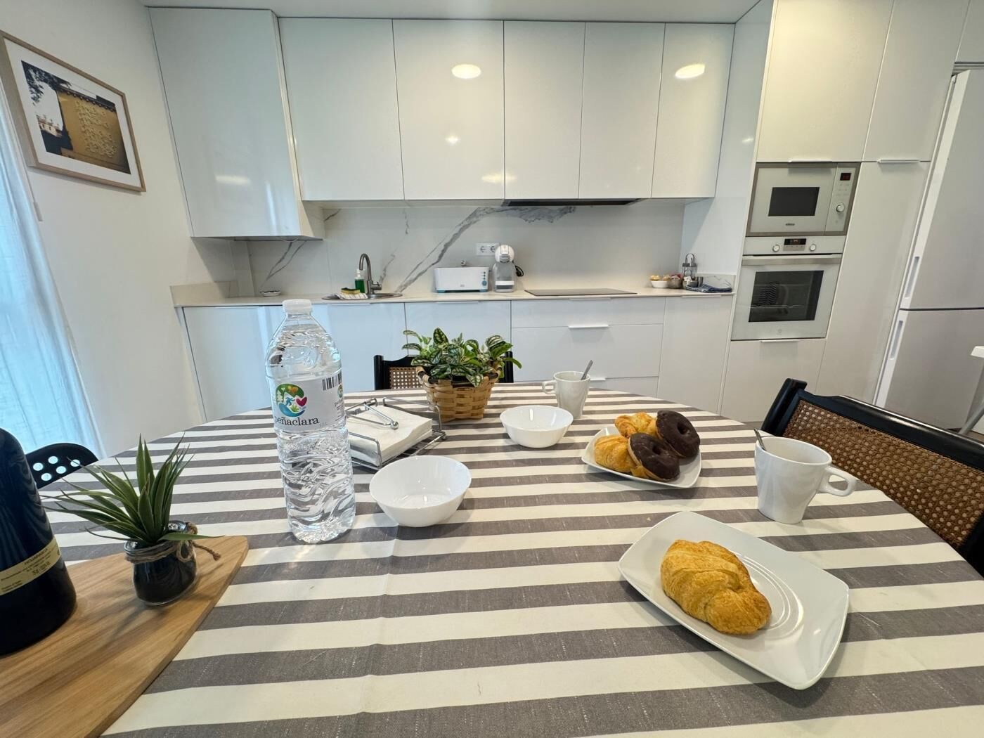 Una cocina moderna con armarios blancos, un mantel a rayas, una botella de agua, un plato de croissants y una taza de café.