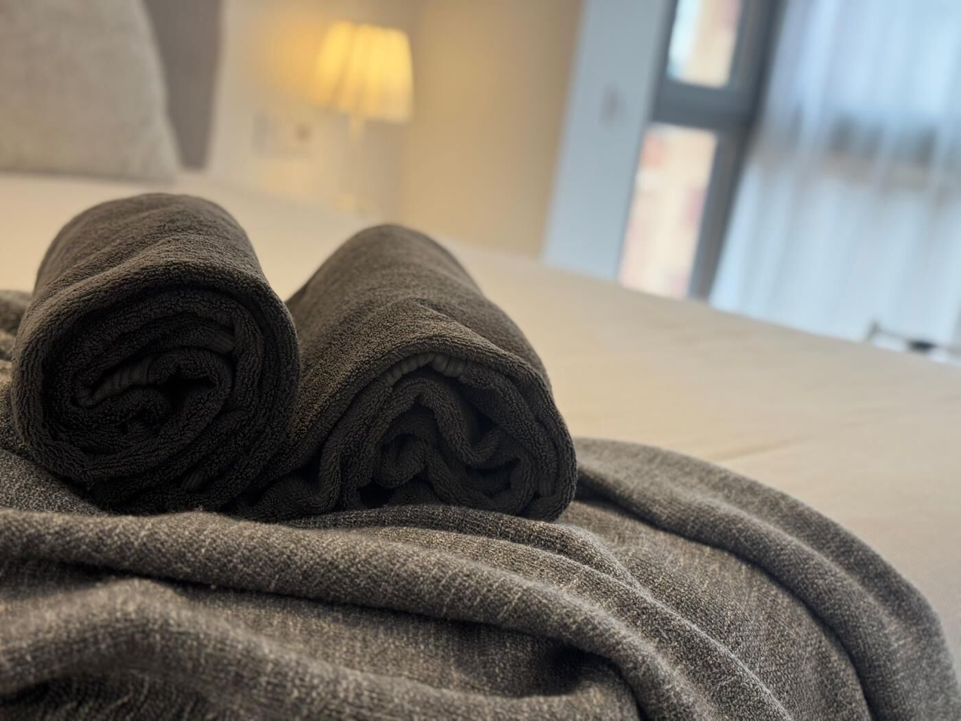 A neatly folded towel on a bed with a blanket, in a room with a lamp and a window.