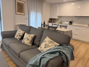 Ein modernes Wohnzimmer mit einem grauen Sofa, gemusterten Kissen, einem Essbereich mit kariertem Tischlein und einer Küche mit weißen Schränken.