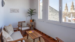 Une pièce chaleureuse avec un plafond en bois, une grande fenêtre donnant sur des bâtiments historiques, une chaise en bois, une petite table avec un livre et une plante en pot.