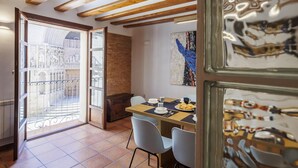 Un coin repas avec une table dressée pour deux personnes, une vue sur une cour intérieure par une porte ouverte et un mur en briques rustiques.