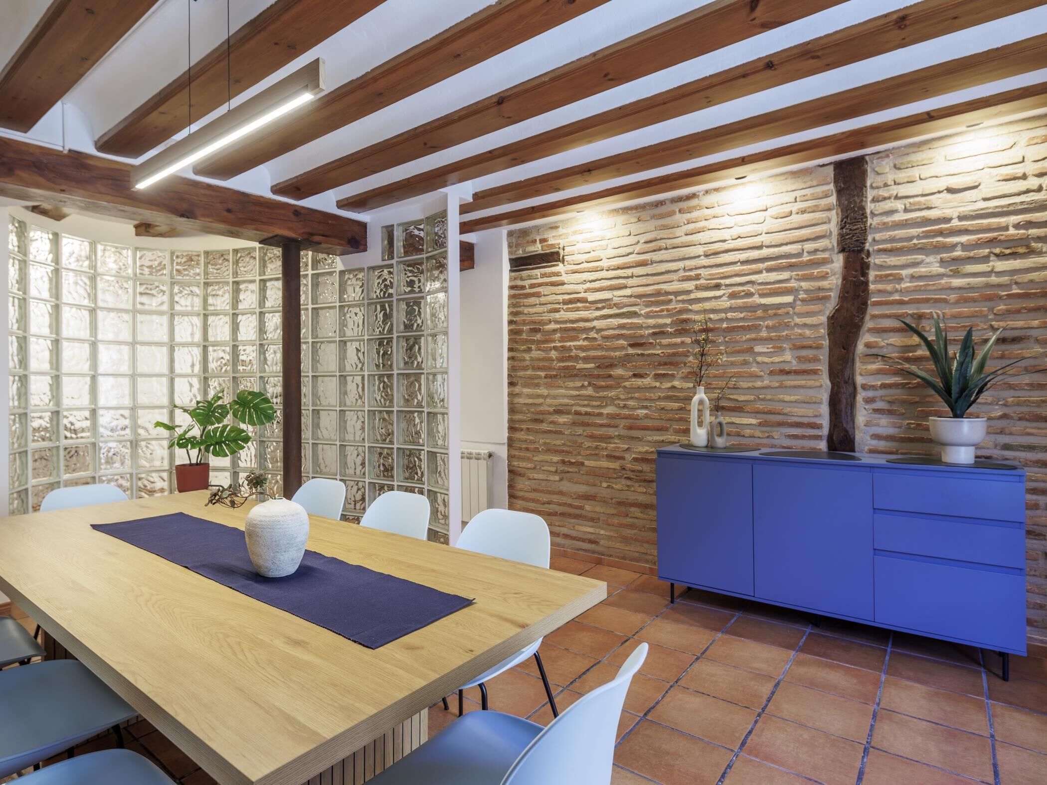 A dining area with a wooden table, white chairs, a blue cabinet, and a brick wall.