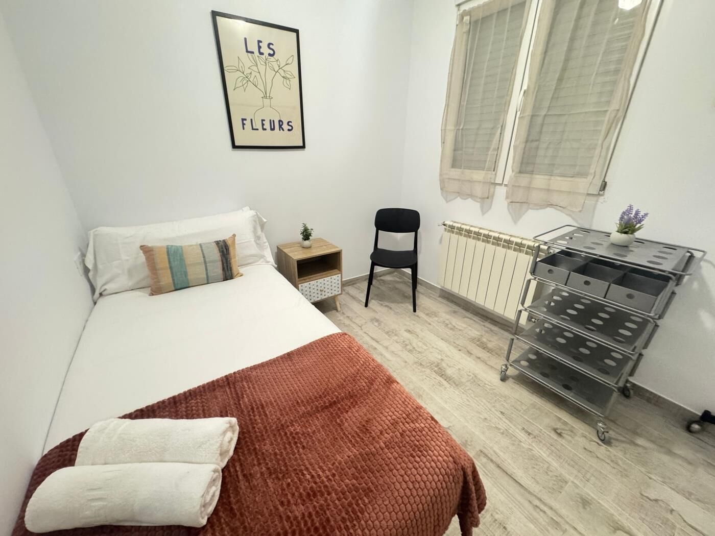 A bedroom with a bed, a chair, a small table with a plant, a radiator, and a wall-mounted picture.