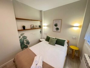 Un dormitorio con cama, mesitas de noche, un taburete pequeño, un estante en la pared con plantas y un cuadro enmarco.