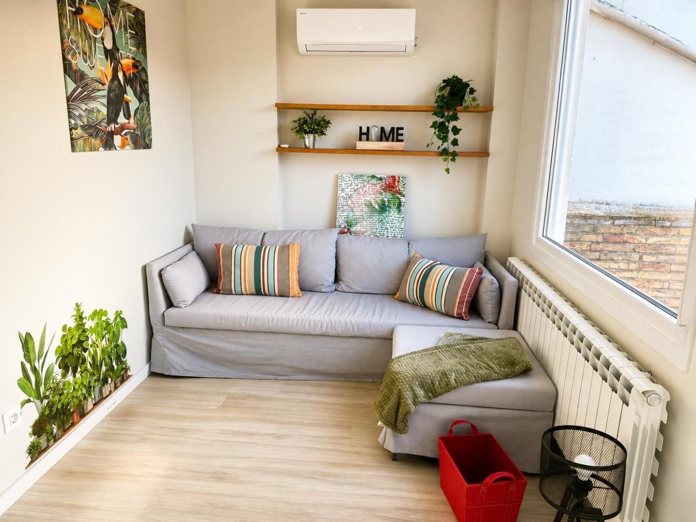 Una sala moderna con un sofá gris, almohadones a rayas, una cesta roja, un radiador y una ventana con vistas.
