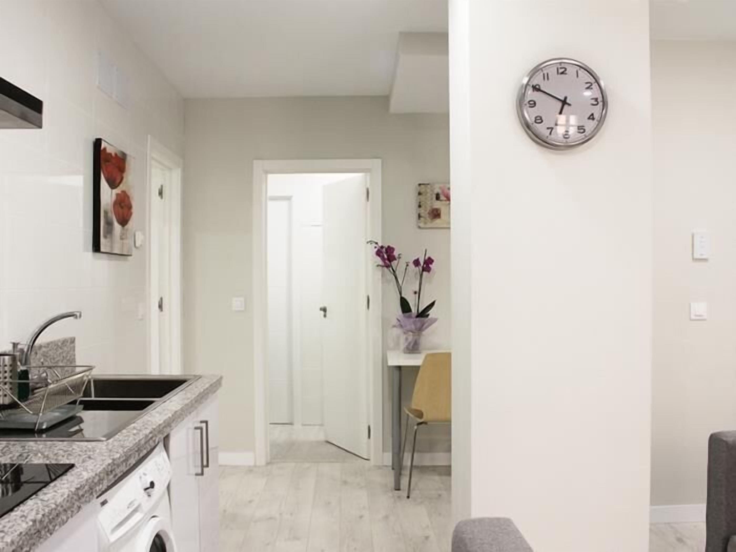 A modern kitchen with a built-in stove, a sink, and a countertop. There is a door leading to another room, and a clock on the wall.