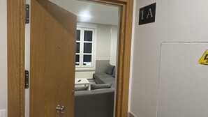 Ein Zimmer mit Holztür, einem grauen Sofa, einem Fenster mit Jalousien und einem Wandzeichen mit '1A'.
