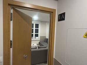 Habitació amb una porta de fusta, un sofà gris i una finestra amb persianes, i un senyal a la paret que diu '1A'.