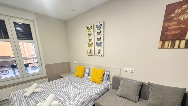 Ein Schlafzimmer mit Bett, grauem Sofa, Fenster mit Jalousien und Wandbildern.
