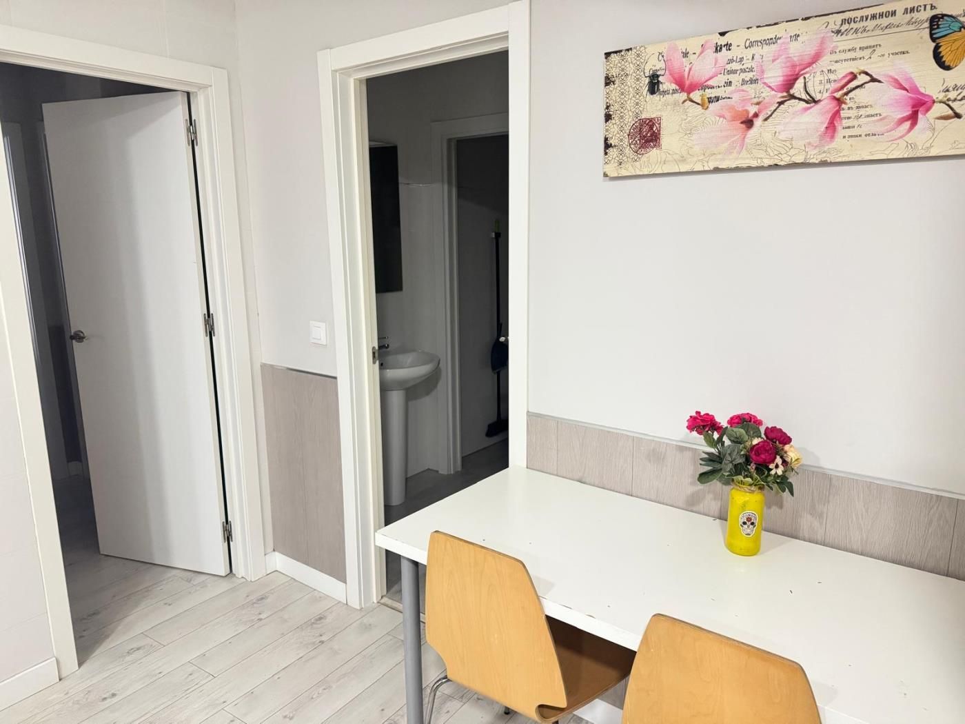 Una habitación con una mesa blanca, dos sillas, un jarrón con flores y un adorno en la pared.