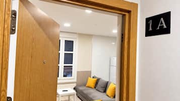 Una habitación con puerta de madera, pared blanca con un letrero negro, sofá gris con cojines amarillos, radiador blanco y una ventana con persianas.