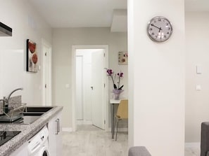 Una cocina moderna con estufa integrada, un lavamanos y una encimera. Hay una puerta que da a otra habitación, y un reloj en la pared.