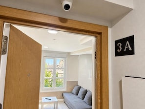 Ein Zimmer mit Holztür, weißem Tisch, grauem Sofa, einem Fenster mit Blick auf Gebäude und einem an der Wand befestigten Schild mit '3A'.