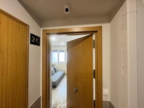 Un couloir avec une porte en bois portant l’inscription « 2C », qui donne sur une chambre à coucher équipée d’un lit et d’une fenêtre.
