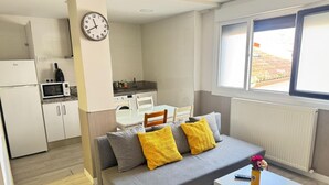 A compact living space with a kitchenette, a sofa with yellow cushions, a dining table, and a clock on the wall. - APARTAMENTOS LOGROÑO CENTRO (LOGROÑO)