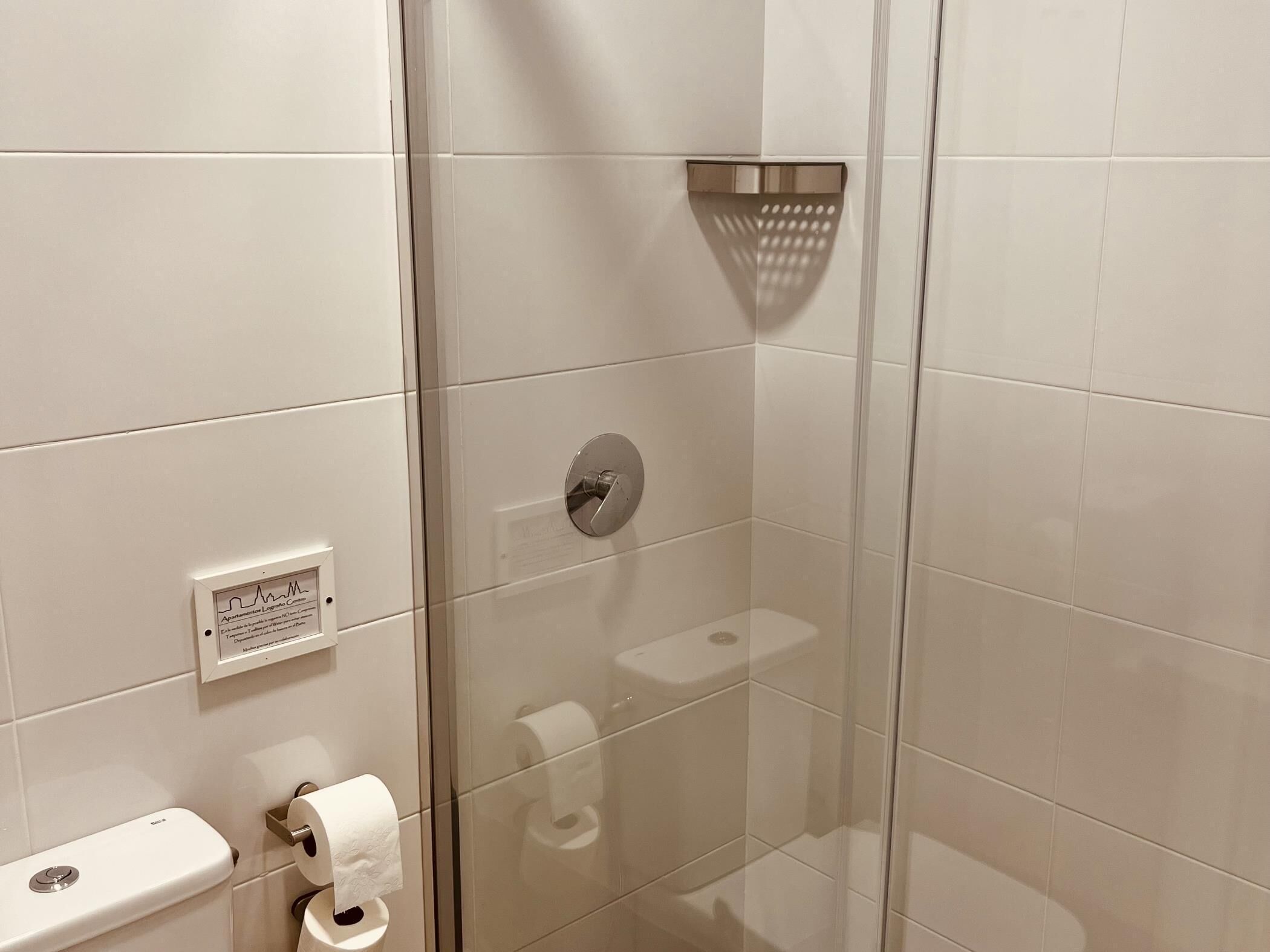 Ein Badezimmer mit weißen Fliesen, einer Duschkabine aus Glas, einem Toilettenpapierhalter und einer sichtbaren Steckdose.