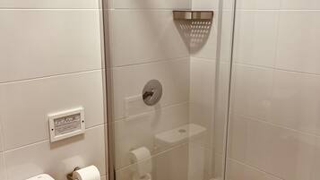 Ein Badezimmer mit weißen Fliesen, einer Duschkabine aus Glas, einem Toilettenpapierhalter und einer sichtbaren Steckdose.