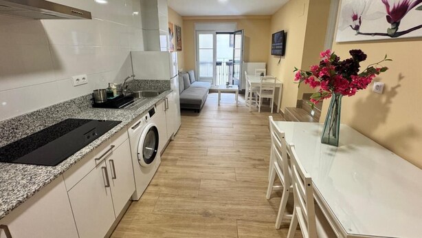A modern kitchen with a built-in washing machine, a dining area with a table and chairs, and a living room with a sofa. - APARTAMENTOS SAN NICOLAS (LOGROÑO)