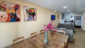 Ein Essbereich mit einem Holztisch, Stühlen und einer Vase mit rosa Blumen. An der Wand hängen zwei Gemälde und im Hintergrund steht ein Fernseher.