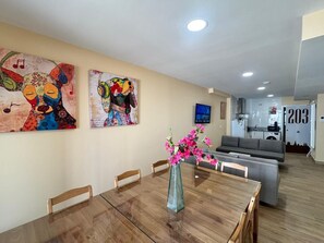 Un coin repas avec une table en bois, des chaises et un vase rempli de fleurs roses. Deux tableaux ornent le mur, et un téléviseur est visible au fond.