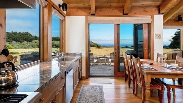 A wooden interior with a kitchen, dining area, and large windows overlooking a beach. - Unit 28 Lot 95 Breaker View (The Sea Ranch)