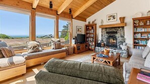A cozy living room with a stone fireplace, wooden bookshelves, and a large window overlooking a scenic view. - Unit 28 Lot 95 Breaker View (The Sea Ranch)