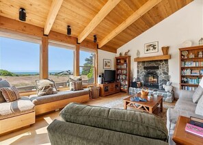 Ein gemütliches Wohnzimmer mit Kamin aus Stein, Holzböschern und einem großen Fenster mit Blick auf eine schöne Landschaft.