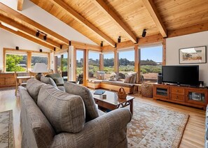Ein großzügiges Wohnzimmer mit einer großen Couch, einem Couchtisch und einem Fernseher. Große Fenster bieten einen Blick ins Freie.