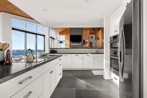 A modern kitchen with white cabinets, a stainless steel refrigerator, and a large window overlooking the ocean.