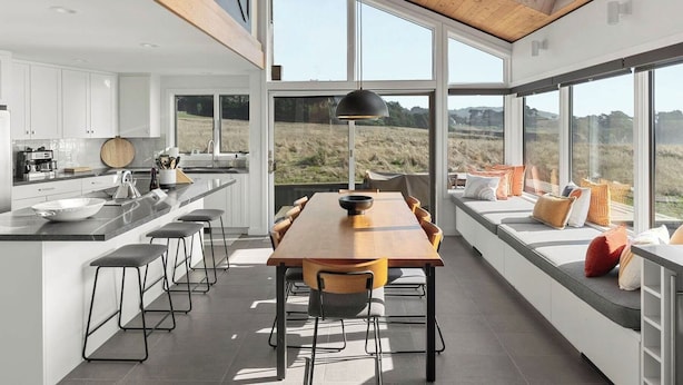 A modern kitchen with a large island, a dining area with a long table, and large windows offering a view of the outdoors.