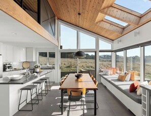 Une cuisine moderne dotée d’un grand îlot central, d’un coin repas avec une longue table et de grandes fenêtres offrant une vue sur l’extérieur.