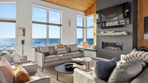 A modern living room with a large window overlooking the ocean, a fireplace, and a flat-screen TV mounted on the wall.