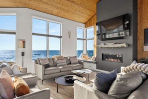 A modern living room with a large window overlooking the ocean, a fireplace, and a flat-screen TV mounted on the wall. - Ocean front bluff top oasis with spectacular views Sleeps up to 8 (The Sea Ranch)