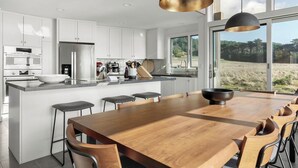 A modern kitchen with white cabinets, a wooden dining table, and a large window offering a view of the outdoors.
