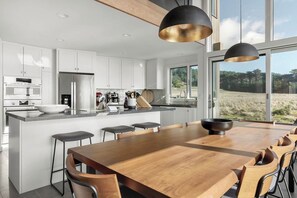 A modern kitchen with white cabinets, a wooden dining table, and a large window offering a view of the outdoors. - Ocean front bluff top oasis with spectacular views Sleeps up to 8 (The Sea Ranch)