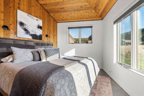 Une chambre avec un plafond en bois, un grand lit avec une literie à motifs, une vue sur l’extérieur par une fenêtre et un tableau accroché au mur.