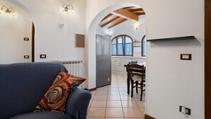 Un soggiorno con divano e cuscino colorato, una cucina con frigorifero e pensili, e un arco che dà accesso a un'altra stanza.