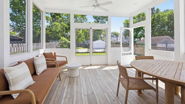 Eine überdachte Terrasse mit Holztisch und -stühlen, einer Couch mit Kissen sowie großen Fenstern mit Blick auf den Garten.