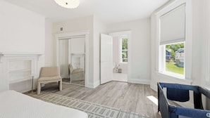 Ein Zimmer mit einem Bett, einem Sessel, einem Fenster mit Jalousien, einer Tür zu einem anderen Raum und einer Deckenleuchte.