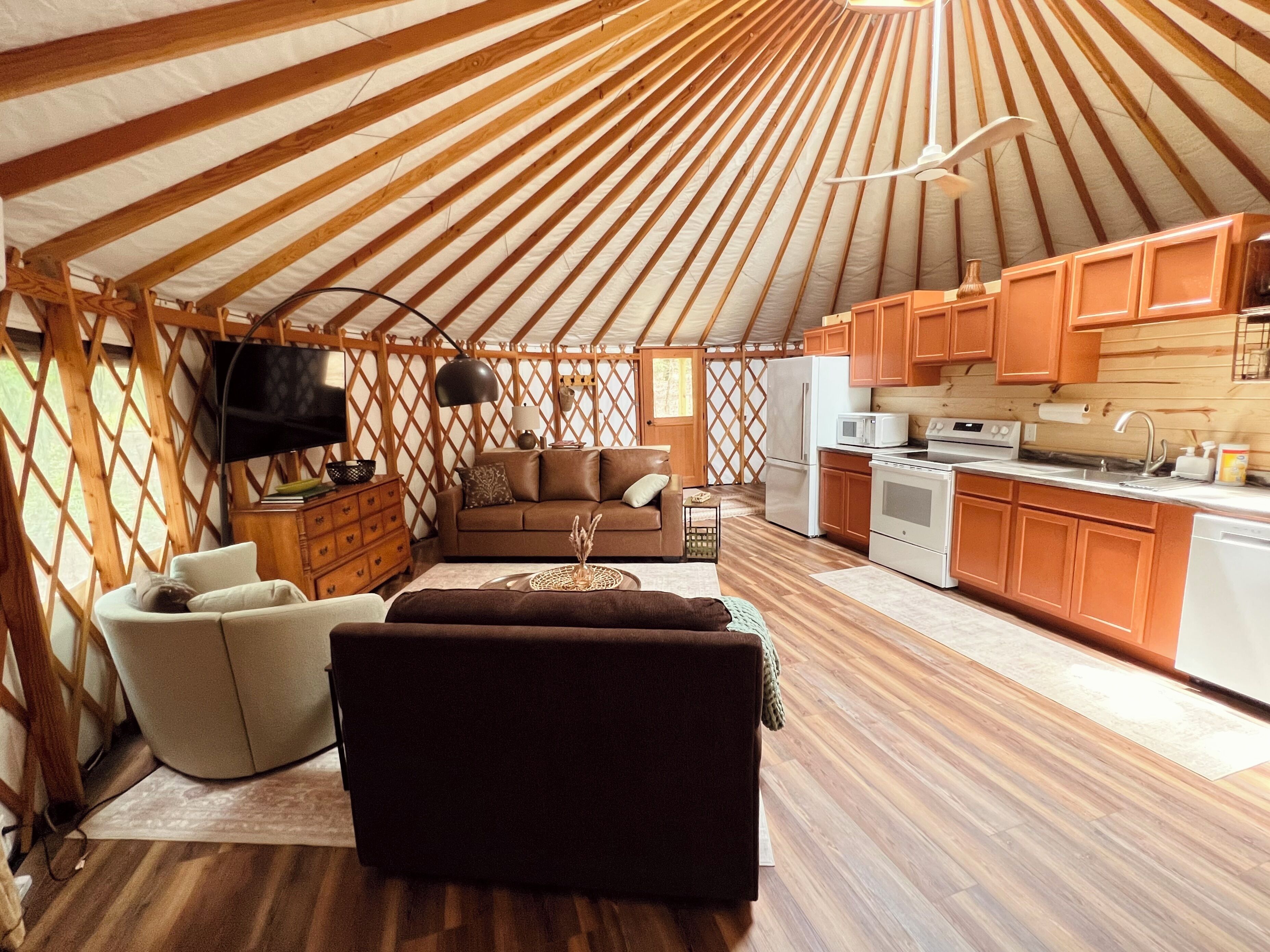 A yurt interior with a sofa, TV, wooden flooring, and a kitchen area.