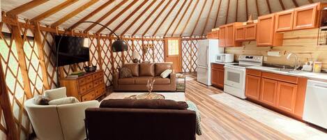 A yurt interior with a sofa, TV, wooden flooring, and a kitchen area.