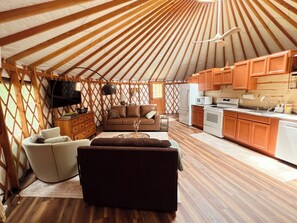 A yurt interior with a sofa, TV, wooden flooring, and a kitchen area. - Luxury Yurt Near Fort Smith Hot Tub, Fishing Pond, & Hiking (Greenwood)