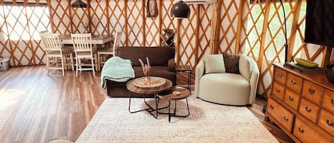 A tented accommodation with a dining area, sofas, a TV, and a wooden cabinet.