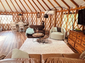 A tented accommodation with a dining area, sofas, a TV, and a wooden cabinet. - Luxury Yurt Near Fort Smith Hot Tub, Fishing Pond, & Hiking (Greenwood)