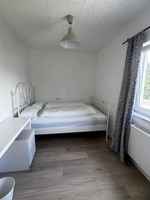 A bedroom with a single bed, a bedside table, a wardrobe, a window with curtains, and a ceiling light.