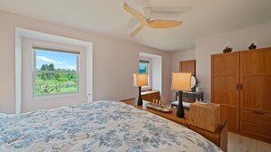 Un dormitorio con una cama grande, ventilador de techo, armario de madera, lámparas de noche y ventana con vistas a la vegetación.