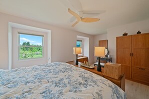 Ein Schlafzimmer mit einem großen Bett, einem Deckenventilator, einem Holzschrank, Nachttischlampen und einem Fenster mit Blick auf Grünflächen.